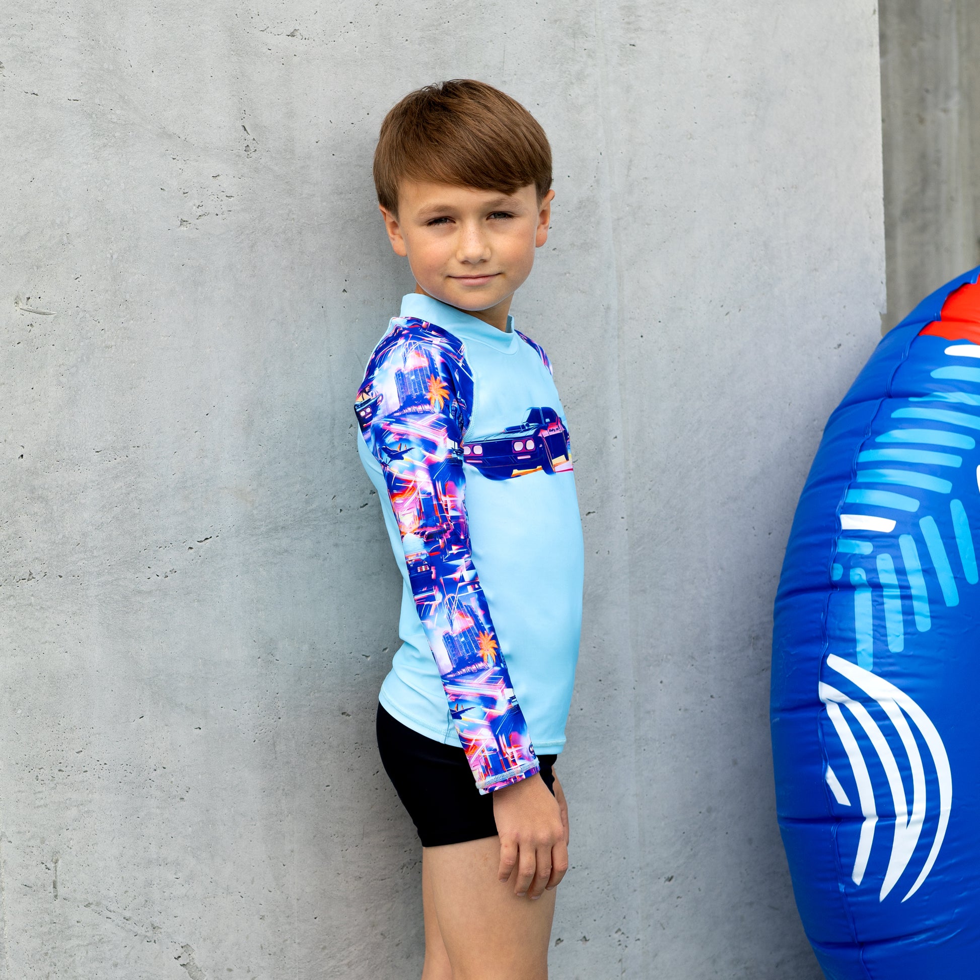 Child wearing a boy long sleeve  rashie with a blue and white inflatable ring next to them against a concrete wall.