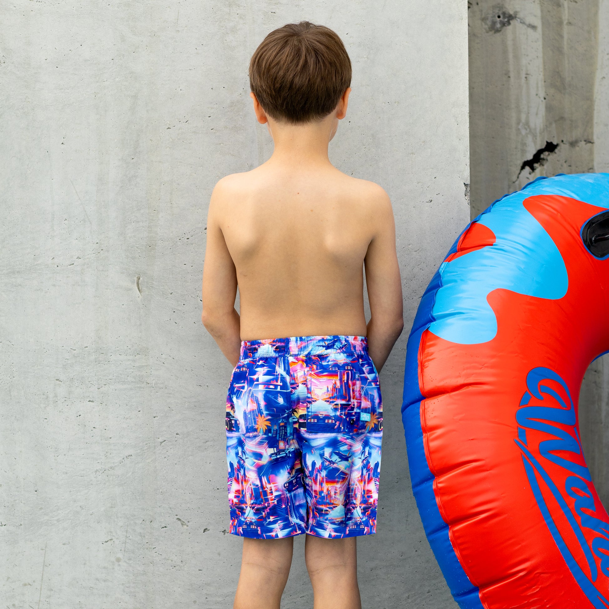 Young boy in colorful boys swim boardies standing next to a red and blue inflatable tube against a gray concrete wall.
