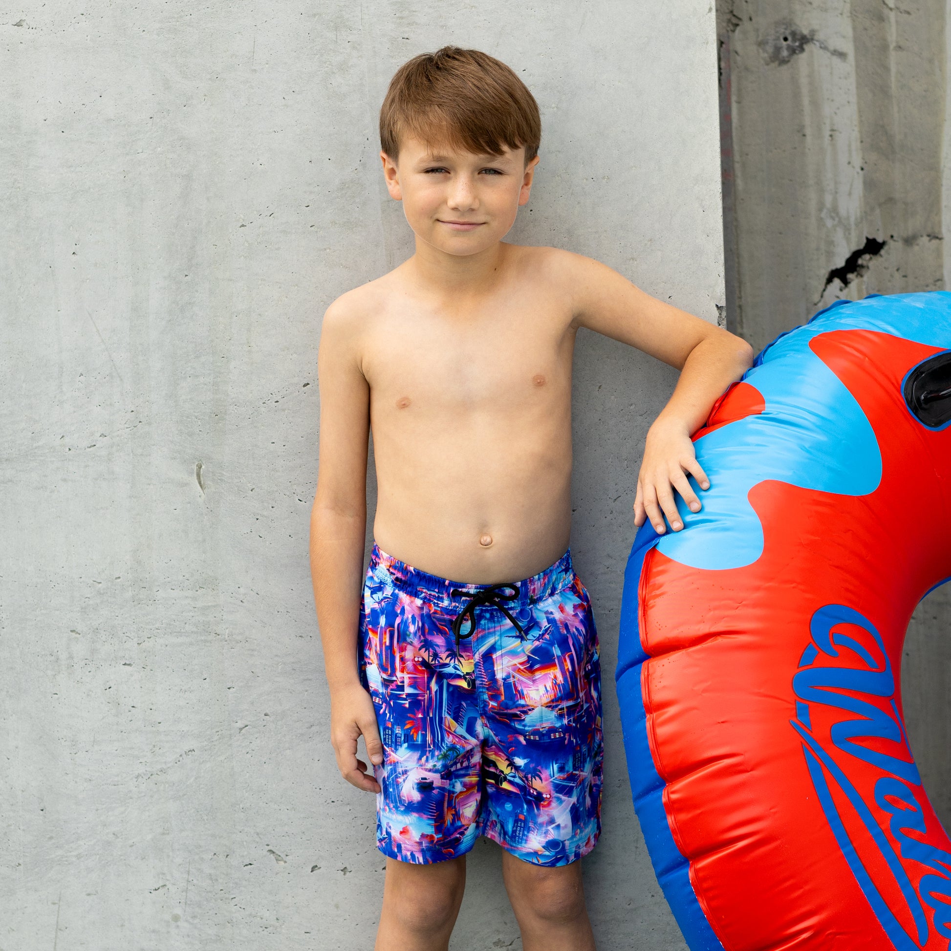 Young boy in colorful boys swim boardies standing next to a red and blue inflatable tube against a gray concrete wall.
