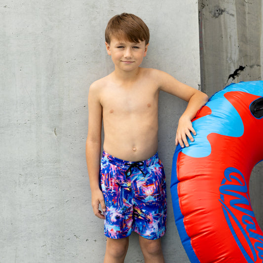 Young boy in colorful boys swim boardies standing next to a red and blue inflatable tube against a gray concrete wall.