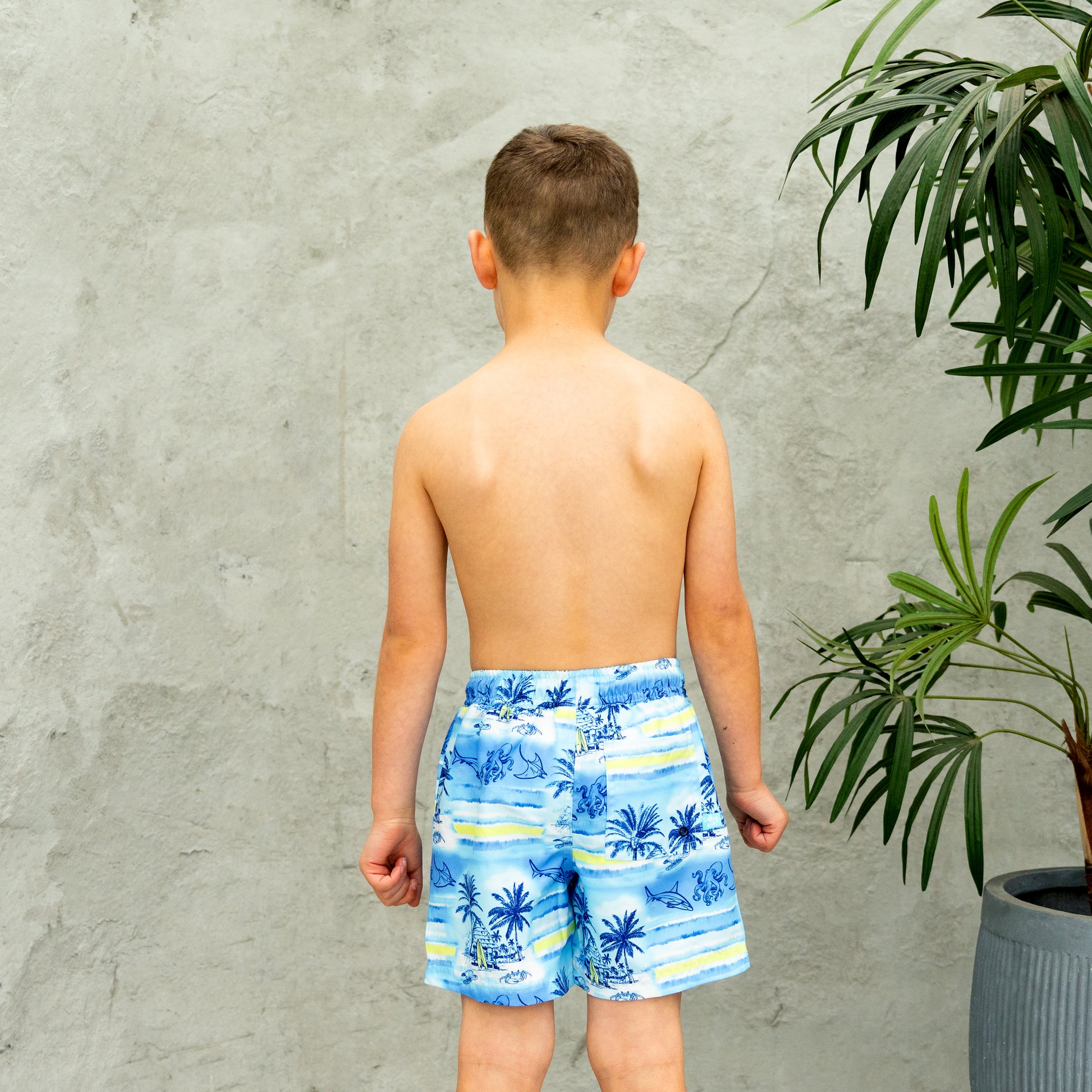 Young boy wearing blue tropical boys swim shorts standing against a gray wall with a plant.