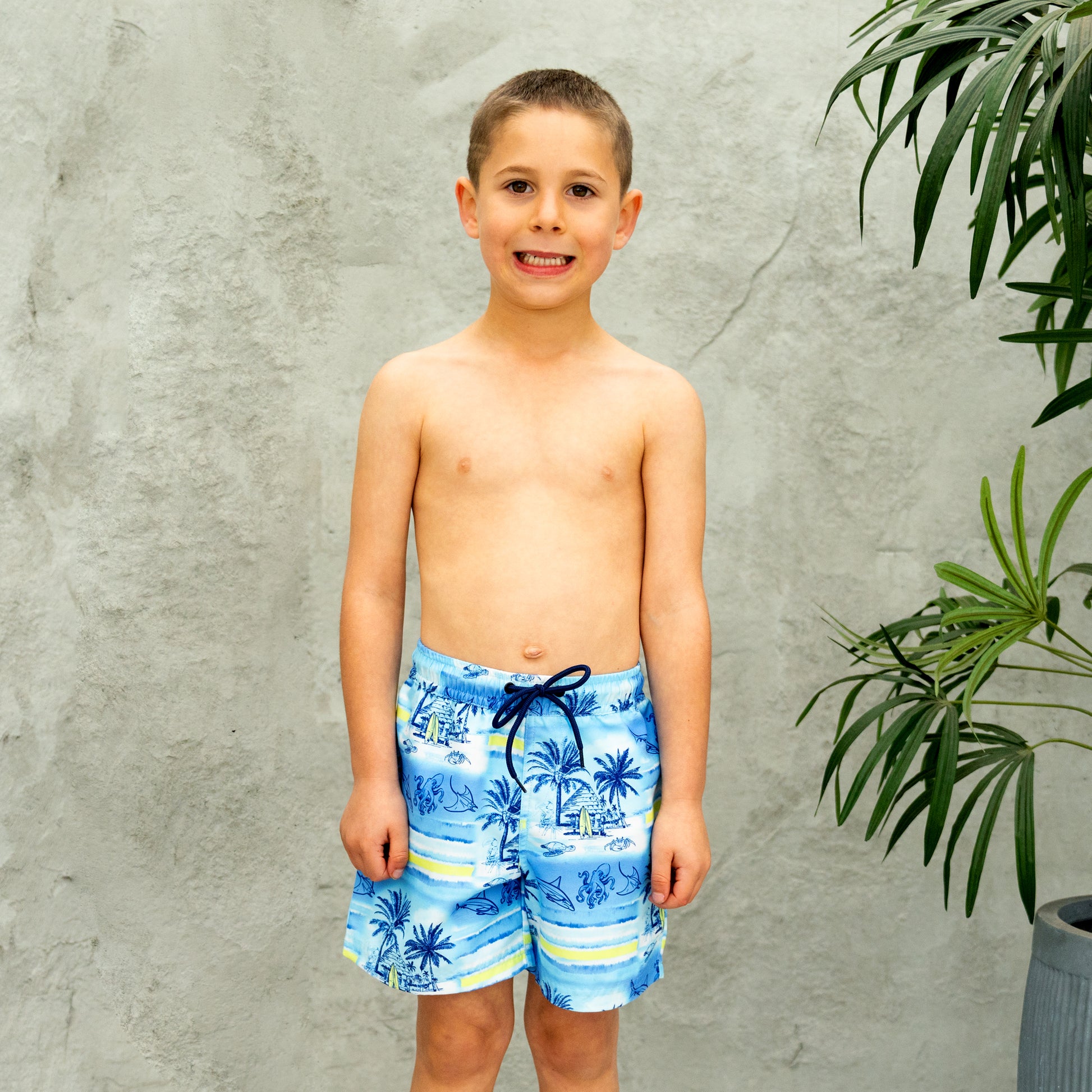 Young boy wearing blue tropical boys swim shorts standing against a gray wall with a plant.