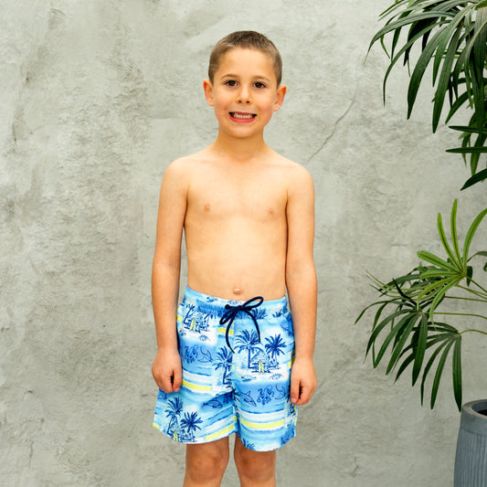 Young boy wearing blue tropical boys swim shorts standing against a gray wall with a plant.