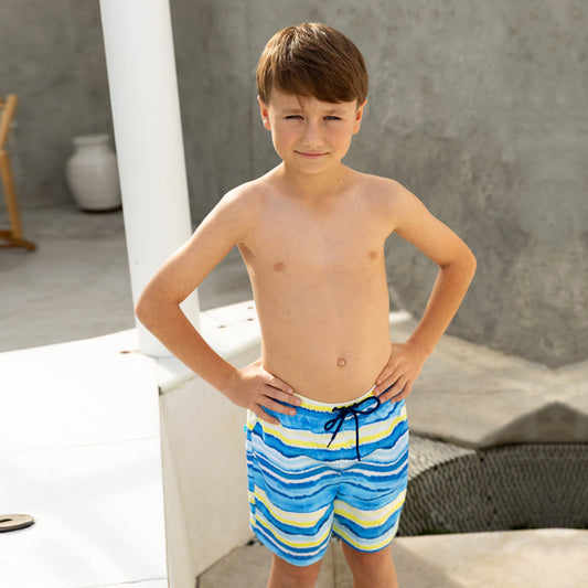 Young boy wearing blue and yellow striped boys swim board shorts standing on a white surface.