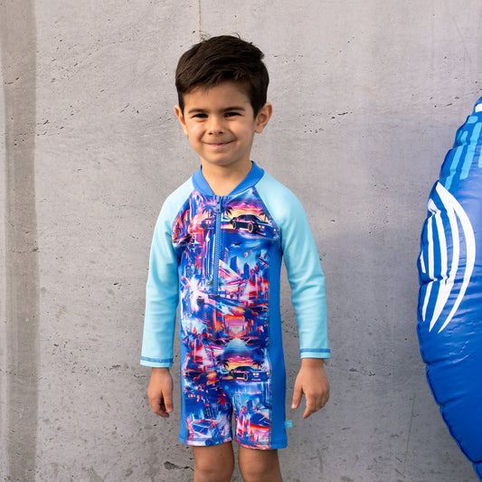 Child wearing a colorful boys all in one swimsuit with blue sleeves against a gray concrete wall