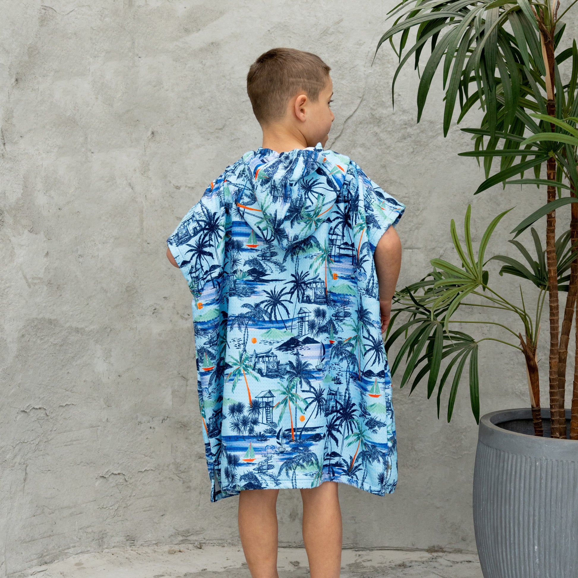 Child wearing a blue tropical print poncho standing against a gray wall with a plant.