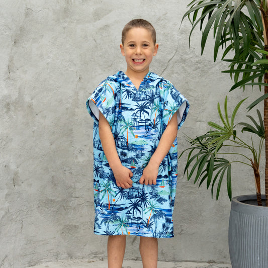 Child wearing a blue tropical patterned poncho standing against a gray wall with a plant.